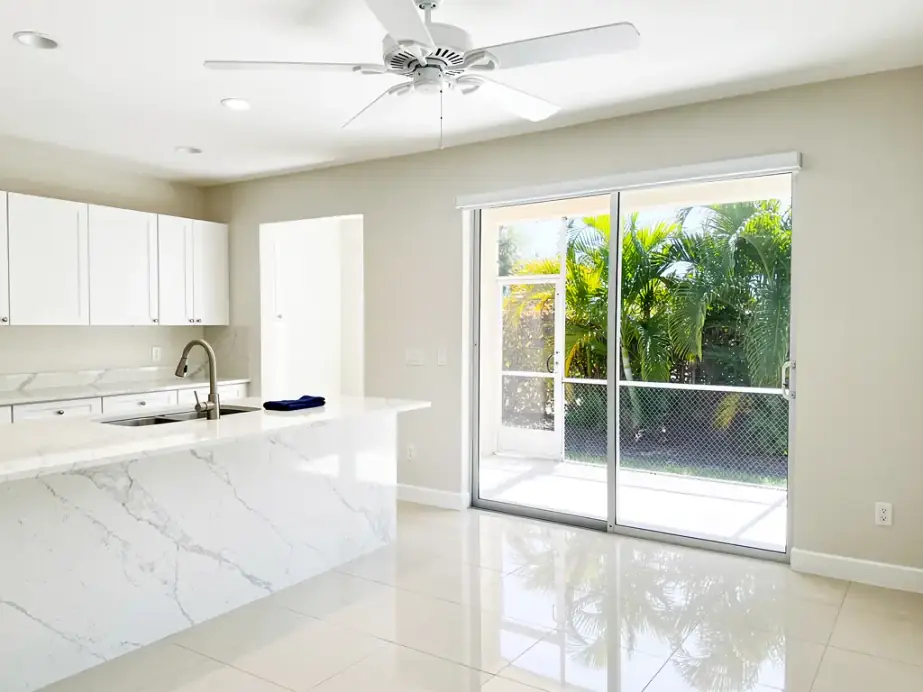 A bright open-plan Florida home after a clean: ceiling fan, white quartz island and sink, glass sliders onto a screened lanai, mirror-sheen tile floor